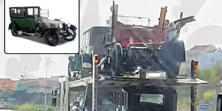 Un vettore trasporta due auto nere d'epoca, tra cui una Rolls-Royce Silver Ghost distrutta al livello superiore, con danni visibili. Un inserto rivela una chiara vista frontale dello stesso modello che guida su una strada alla luce del giorno.