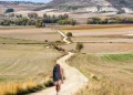 Silvana Di Liberto, di spalle, con un grande zaino cammina da sola sulla Via de la Plata, seguendo un tortuoso sentiero sterrato attraverso campi aperti verso un villaggio lontano, circondato da dolci colline e terreni agricoli a macchia d'olio sotto un cielo parzialmente nuvoloso.