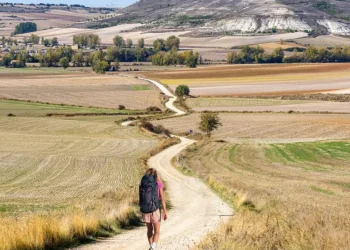 Silvana Di Liberto, di spalle, con un grande zaino cammina da sola sulla Via de la Plata, seguendo un tortuoso sentiero sterrato attraverso campi aperti verso un villaggio lontano, circondato da dolci colline e terreni agricoli a macchia d'olio sotto un cielo parzialmente nuvoloso.