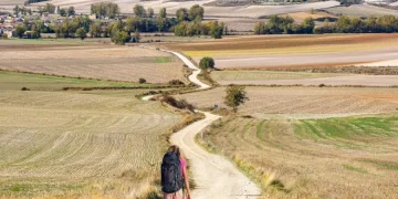 Silvana Di Liberto, di spalle, con un grande zaino cammina da sola sulla Via de la Plata, seguendo un tortuoso sentiero sterrato attraverso campi aperti verso un villaggio lontano, circondato da dolci colline e terreni agricoli a macchia d'olio sotto un cielo parzialmente nuvoloso.