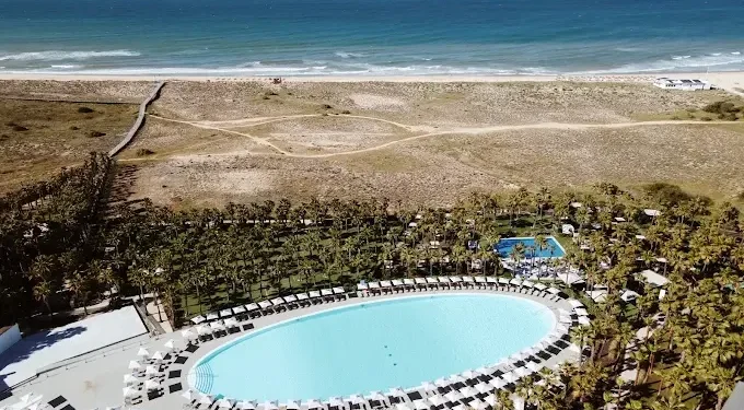 Una grande piscina ovale circondata da sedie a sdraio e palme al Wine & Books Algarve, situato vicino a una spiaggia sabbiosa e all'oceano sotto un cielo limpido. Dall'area del resort partono sentieri che conducono alla spiaggia.