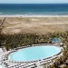 Una grande piscina ovale circondata da sedie a sdraio e palme al Wine & Books Algarve, situato vicino a una spiaggia sabbiosa e all'oceano sotto un cielo limpido. Dall'area del resort partono sentieri che conducono alla spiaggia.