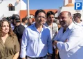 Rui Cristina eletto indaco di Albufeira con gruppo di persone all'aperto in una giornata di sole, con un uomo in camicia bianca e una donna in abito color oliva in testa, riuniti per festeggiare i risultati delle elezioni amministrative di Albufeira. Sullo sfondo sono visibili edifici bianchi e un cartello di parcheggio blu.