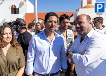 Rui Cristina eletto indaco di Albufeira con gruppo di persone all'aperto in una giornata di sole, con un uomo in camicia bianca e una donna in abito color oliva in testa, riuniti per festeggiare i risultati delle elezioni amministrative di Albufeira. Sullo sfondo sono visibili edifici bianchi e un cartello di parcheggio blu.