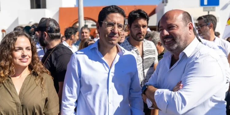 Rui Cristina eletto indaco di Albufeira con gruppo di persone all'aperto in una giornata di sole, con un uomo in camicia bianca e una donna in abito color oliva in testa, riuniti per festeggiare i risultati delle elezioni amministrative di Albufeira. Sullo sfondo sono visibili edifici bianchi e un cartello di parcheggio blu.