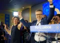 Carlos Moedas e una donna festeggiano i risultati delle elezioni amministrative di Lisbona sul palco, alzando le mani in segno di vittoria. Si trovano su un podio con un cartello "Lisboa 2025" e sorridendo ai sostenitori, mentre le persone applaudono nella sala illuminata.