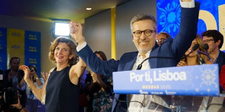 Carlos Moedas e una donna festeggiano i risultati delle elezioni amministrative di Lisbona sul palco, alzando le mani in segno di vittoria. Si trovano su un podio con un cartello "Lisboa 2025" e sorridendo ai sostenitori, mentre le persone applaudono nella sala illuminata.