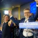 Carlos Moedas e una donna festeggiano i risultati delle elezioni amministrative di Lisbona sul palco, alzando le mani in segno di vittoria. Si trovano su un podio con un cartello "Lisboa 2025" e sorridendo ai sostenitori, mentre le persone applaudono nella sala illuminata.