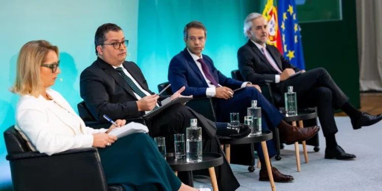 Quattro persone tra cui il ministro dell'economia Miranda Sarmento siedono su un palco con microfoni e documenti per la presentazione della legge finanziaria portoghese 2026, davanti a uno sfondo verde con bandiere portoghesi e dell'UE. Sui tavoli accanto a loro c'è dell'acqua in bottiglia.