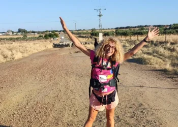 Silvana Di Liberto, con le braccia alzate, con una maglietta rosa, sorride ed è sola sulla Via de la Plata, seguendo un tortuoso sentiero sterrato attraverso campi aperti verso un villaggio lontano, circondato da dolci colline e terreni agricoli a macchia d'olio sotto un cielo parzialmente nuvoloso.