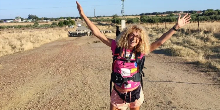 Silvana Di Liberto, con le braccia alzate, con una maglietta rosa, sorride ed è sola sulla Via de la Plata, seguendo un tortuoso sentiero sterrato attraverso campi aperti verso un villaggio lontano, circondato da dolci colline e terreni agricoli a macchia d'olio sotto un cielo parzialmente nuvoloso.