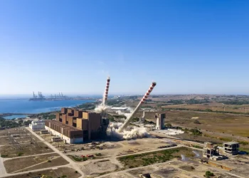 Frame del video di demolizione in veduta aerea di due alte ciminiere di una centrale elettrica, che si appoggiano e crollano tra nuvole di polvere vicino a un edificio industriale sulla costa, con un cielo azzurro e gru portuali lontane sullo sfondo.