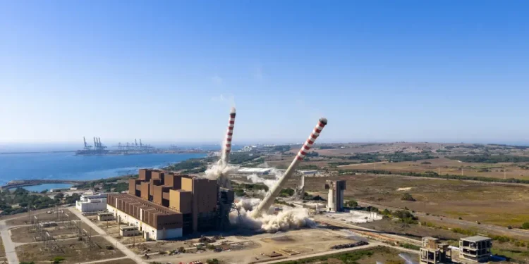 Frame del video di demolizione in veduta aerea di due alte ciminiere di una centrale elettrica, che si appoggiano e crollano tra nuvole di polvere vicino a un edificio industriale sulla costa, con un cielo azzurro e gru portuali lontane sullo sfondo.