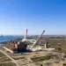Frame del video di demolizione in veduta aerea di due alte ciminiere di una centrale elettrica, che si appoggiano e crollano tra nuvole di polvere vicino a un edificio industriale sulla costa, con un cielo azzurro e gru portuali lontane sullo sfondo.