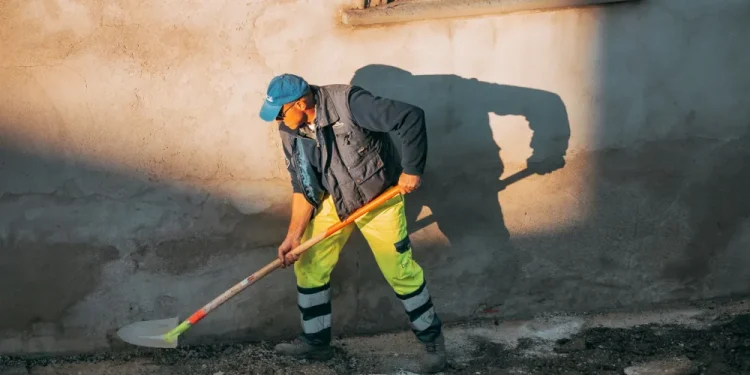 Un operaio edile con berretto, gilet e pantaloni a righe gialle usa una pala per scavare in una strada illuminata dal sole, illustrando il costante andamento dell' economia portoghese accanto a un muro beige con una finestra che proietta ombre.