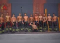 Un gruppo di undici vigili del fuoco a torso nudo, presenti nel Calendario vigili del fuoco portoghesi, posano davanti a un edificio, indossando pantaloni e stivali da pompiere, tenendo in mano vari attrezzi, con uno inginocchiato di fronte a loro e che si flette.