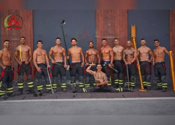 Un gruppo di undici vigili del fuoco a torso nudo, presenti nel Calendario vigili del fuoco portoghesi, posano davanti a un edificio, indossando pantaloni e stivali da pompiere, tenendo in mano vari attrezzi, con uno inginocchiato di fronte a loro e che si flette.
