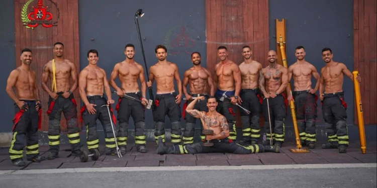 Un gruppo di undici vigili del fuoco a torso nudo, presenti nel Calendario vigili del fuoco portoghesi, posano davanti a un edificio, indossando pantaloni e stivali da pompiere, tenendo in mano vari attrezzi, con uno inginocchiato di fronte a loro e che si flette.