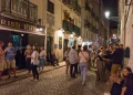 Una folla vivace si raduna di notte fuori da un pub irlandese su una strada acciottolata del Bairro Alto di Lisbona; le persone chiacchierano, stanno in piedi e si siedono, mentre gli edifici illuminati dai lampioni vantano balconi e bandiere, spesso poi restano scene di degrado.
