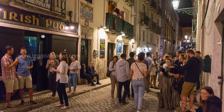 Una folla vivace si raduna di notte fuori da un pub irlandese su una strada acciottolata del Bairro Alto di Lisbona; le persone chiacchierano, stanno in piedi e si siedono, mentre gli edifici illuminati dai lampioni vantano balconi e bandiere, spesso poi restano scene di degrado.