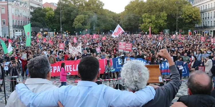 Quattro persone con le braccia al collo affrontano una grande folla durante una protesta all'aperto o una manifestazione contro il Portogallo contro la riforma del lavoro, mentre molti tra la folla reggono striscioni e cartelli colorati, con alberi ed edifici sullo sfondo.