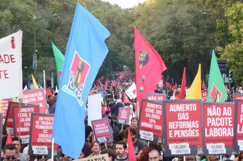 Una grande folla durante una protesta all'aperto o una manifestazione nelle piazze in Portogallo contro la riforma del lavoro, mentre molti tra la folla reggono striscioni e cartelli colorati, con alberi ed edifici sullo sfondo.