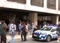 Una folla di persone attende fuori da un edificio con grandi finestre in quella che sembra essere una protesta contro il Portogallo negativo per l'immigrazione, mentre un'auto della polizia con luci lampeggianti è parcheggiata di fronte. Diverse persone sono in piedi o camminano sul marciapiede.
