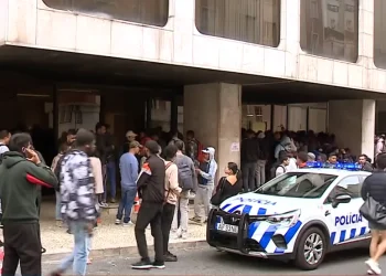 Una folla di persone attende fuori da un edificio con grandi finestre in quella che sembra essere una protesta contro il Portogallo negativo per l'immigrazione, mentre un'auto della polizia con luci lampeggianti è parcheggiata di fronte. Diverse persone sono in piedi o camminano sul marciapiede.