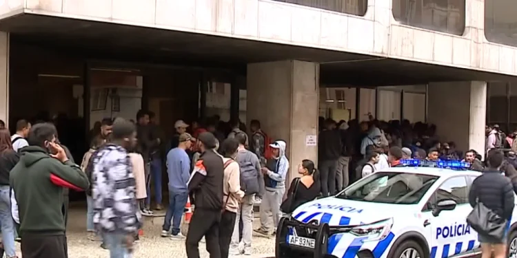 Una folla di persone attende fuori da un edificio con grandi finestre in quella che sembra essere una protesta contro il Portogallo negativo per l'immigrazione, mentre un'auto della polizia con luci lampeggianti è parcheggiata di fronte. Diverse persone sono in piedi o camminano sul marciapiede.
