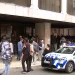 Una folla di persone attende fuori da un edificio con grandi finestre in quella che sembra essere una protesta contro il Portogallo negativo per l'immigrazione, mentre un'auto della polizia con luci lampeggianti è parcheggiata di fronte. Diverse persone sono in piedi o camminano sul marciapiede.