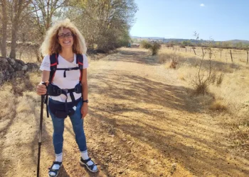 Silvana Di Liberto con i capelli ricci e gli occhiali si accovaccia e sorride all'obiettivo su un sentiero lungo la Via de la Plata, in Castilla y Leon. Sullo sfondo, alcuni alberi, delle colline e un cielo azzurro.