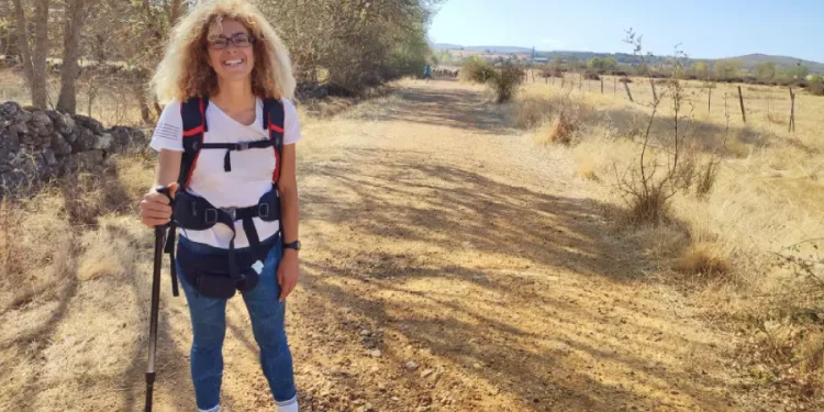 Silvana Di Liberto con i capelli ricci e gli occhiali si accovaccia e sorride all'obiettivo su un sentiero lungo la Via de la Plata, in Castilla y Leon. Sullo sfondo, alcuni alberi, delle colline e un cielo azzurro.