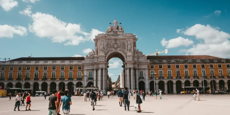 La gente cammina in piazza do Comercio di Lisbona, sullo sfondo del grande arco di pietra, fiancheggiato da edifici gialli sotto un cielo blu con nuvole sparse: una scena vivace che riflette l'alto indice di benessere per cui Portogallo è conosciuta.