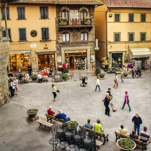 La piazza di una vivace città in Italia riflette l'alto indice di benessere, con persone che passeggiano, si siedono sulle panchine e socializzano. Circondata da edifici storici, negozi, caffè con fioriere sui balconi e posti a sedere all'aperto punteggiati da piante in vaso.