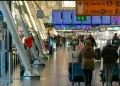 Un affollato terminal aeroportuale con viaggiatori, tra cui italiani diretti all'estero, che camminano e tirano i bagagli osservando i dati info del 2024. Alcuni sono seduti su panchine mentre i cartelli mostrano le indicazioni per il check-in e i gate. L'area è luminosa con luce naturale proveniente da grandi finestre.