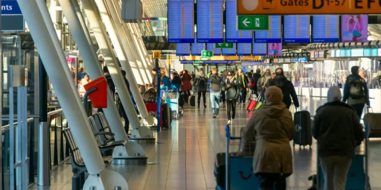 Un affollato terminal aeroportuale con viaggiatori, tra cui italiani diretti all'estero, che camminano e tirano i bagagli osservando i dati info del 2024. Alcuni sono seduti su panchine mentre i cartelli mostrano le indicazioni per il check-in e i gate. L'area è luminosa con luce naturale proveniente da grandi finestre.