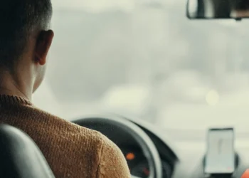 Una persona che indossa un maglione marrone è alla guida di un'auto, vista dal sedile posteriore. Sul cruscotto è montato uno smartphone con un'applicazione di navigazione e il brevetto digitale Portogallo. La vista esterna attraverso il parabrezza è sfocata.