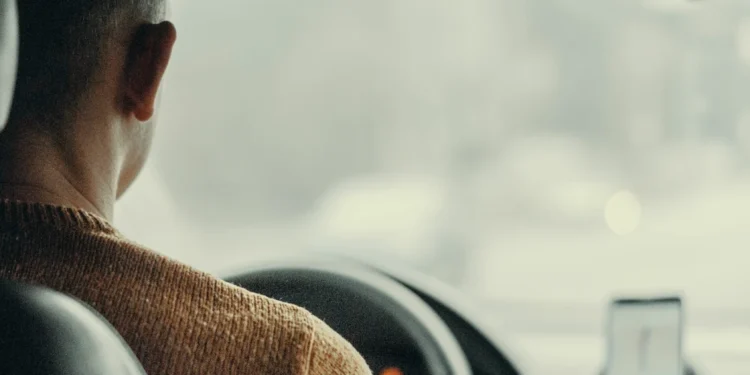 Una persona che indossa un maglione marrone è alla guida di un'auto, vista dal sedile posteriore. Sul cruscotto è montato uno smartphone con un'applicazione di navigazione e il brevetto digitale Portogallo. La vista esterna attraverso il parabrezza è sfocata.