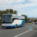 Un pullman bianco e blu, percorre in sicurezza un'autostrada in Portogallo con alberi e colline sullo sfondo. I cartelli stradali indicano Algarve e Lisbona sotto un cielo parzialmente nuvoloso.