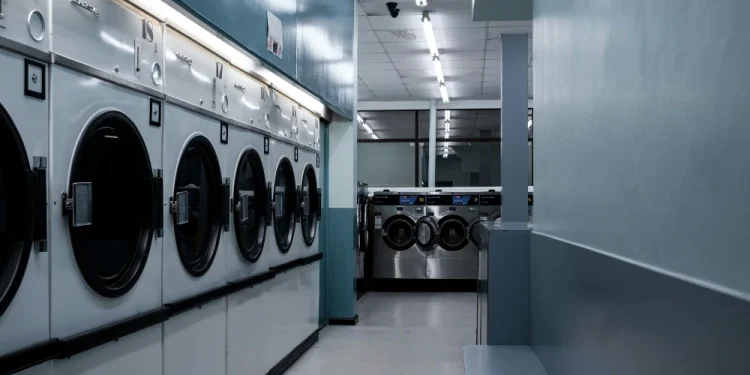 Una fila di grandi lavatrici a carica frontale si estende sul lato sinistro di una lavanderia a gettoni vuota e pulita, la Coin Laundry Portogallo, con un'illuminazione fluorescente e un pavimento lucido. Altre macchine sono visibili sullo sfondo.