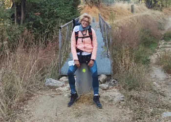 Silvana Di Liberto con giacca e zaino rosa sorride seduta su una roccia lungo la via Plata nella Estremadura, davanti a un piccolo ponte di legno circondato da erba secca e alberi.