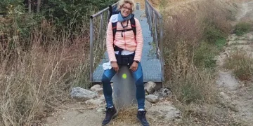 Silvana Di Liberto con giacca e zaino rosa sorride seduta su una roccia lungo la via Plata nella Estremadura, davanti a un piccolo ponte di legno circondato da erba secca e alberi.