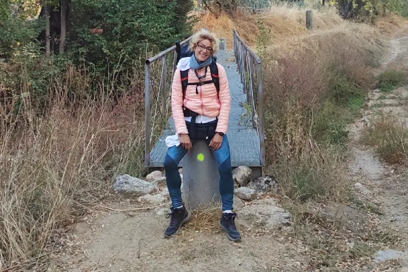 Silvana Di Liberto con giacca e zaino rosa sorride seduta su una roccia lungo la via Plata nella Estremadura, davanti a un piccolo ponte di legno circondato da erba secca e alberi.