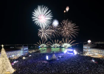 Una grande folla si raduna in piazza a Lisbona di notte per il Capodanno, osservando in sicurezza i vibranti fuochi d'artificio sull'acqua. Gli edifici brillano e un gigantesco albero di Natale scintilla di luci sulla sinistra, con misure di sicurezza rafforzate.