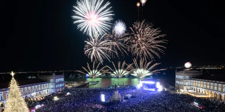 Una grande folla si raduna in piazza a Lisbona di notte per il Capodanno, osservando in sicurezza i vibranti fuochi d'artificio sull'acqua. Gli edifici brillano e un gigantesco albero di Natale scintilla di luci sulla sinistra, con misure di sicurezza rafforzate.