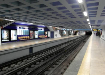 La stazione della metropolitana di Santa Apolónia, prima della chiusuara, pulita e moderna, con banchine vuote, pavimenti piastrellati e manifesti pubblicitari alle pareti. I binari corrono al centro, mentre le luminose luci del soffitto illuminano l'ampia area.