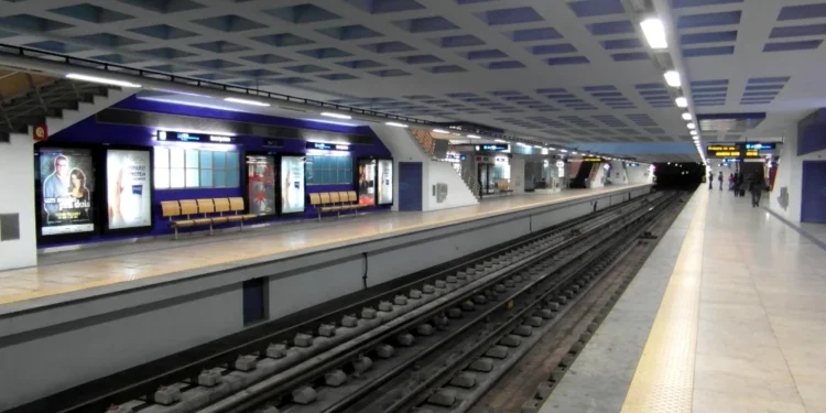 La stazione della metropolitana di Santa Apolónia, prima della chiusuara, pulita e moderna, con banchine vuote, pavimenti piastrellati e manifesti pubblicitari alle pareti. I binari corrono al centro, mentre le luminose luci del soffitto illuminano l'ampia area.