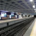 La stazione della metropolitana di Santa Apolónia, prima della chiusuara, pulita e moderna, con banchine vuote, pavimenti piastrellati e manifesti pubblicitari alle pareti. I binari corrono al centro, mentre le luminose luci del soffitto illuminano l'ampia area.