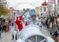Babbo Natale e una persona con un costume d'argento lucido viaggiano su un'auto bianca durante la parata di Natale a Loulé in Algarve, circondati dalla folla e da bambini che guardano e scattano foto. Decorazioni ed edifici fanno da sfondo.