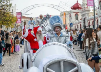 Babbo Natale e una persona con un costume d'argento lucido viaggiano su un'auto bianca durante la parata di Natale a Loulé in Algarve, circondati dalla folla e da bambini che guardano e scattano foto. Decorazioni ed edifici fanno da sfondo.